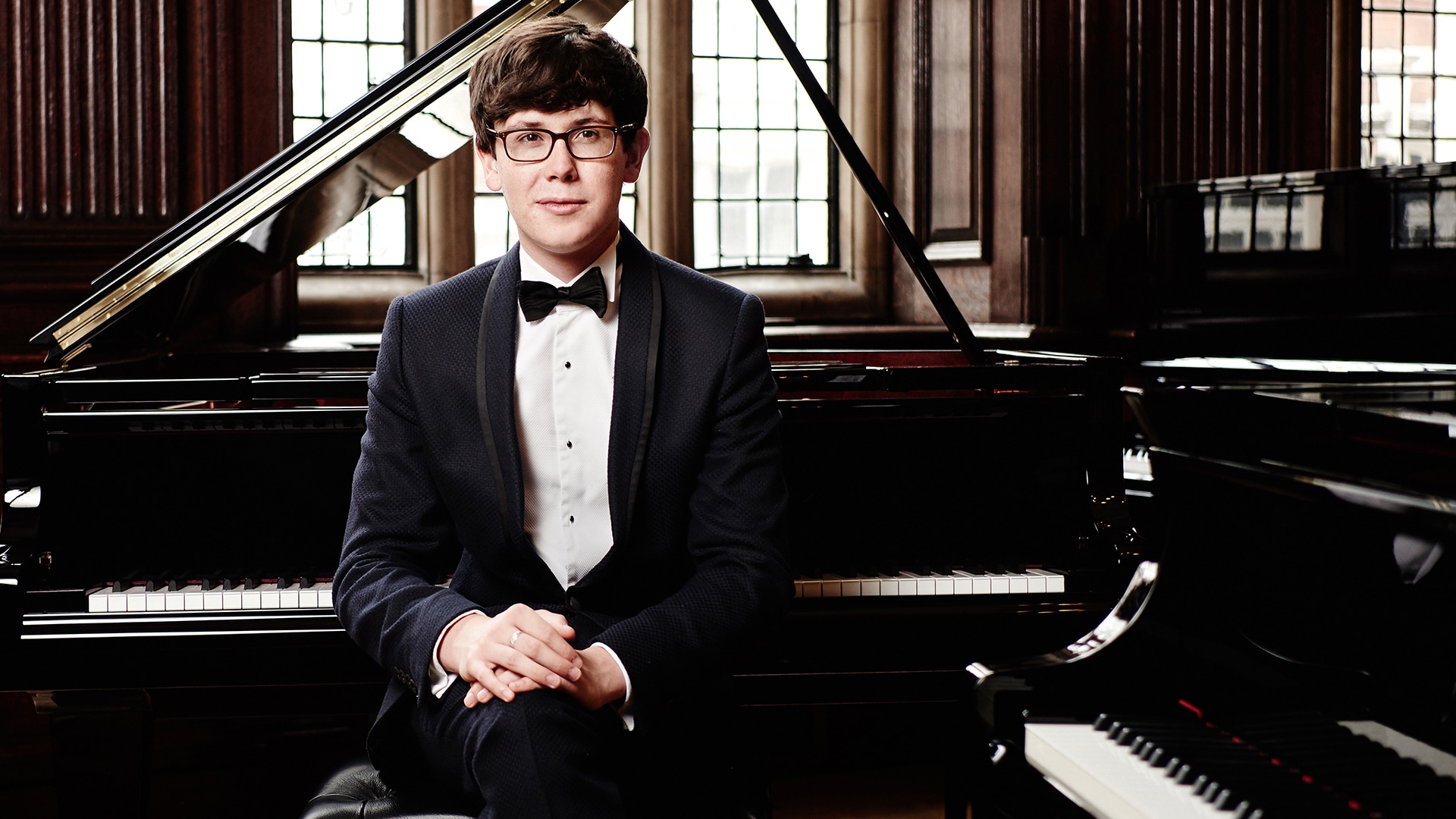 Martin James Bartlett wearing a suit and bow tie, sat on a piano stool in front of a grand piano. Martin wears glasses and has short, wavy brown hair.
