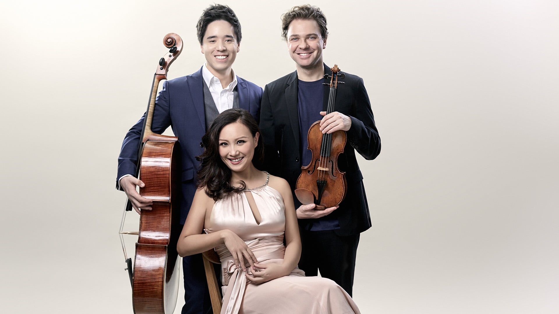 The Sitkovetsky Trio. Two stood wearing blazers, both with short brown hair, one holding a cello, one holding a violin. One sat on a chair in front of them with curly brown shoulder length hair, wearing a formal rose-coloured gown. All smiling.