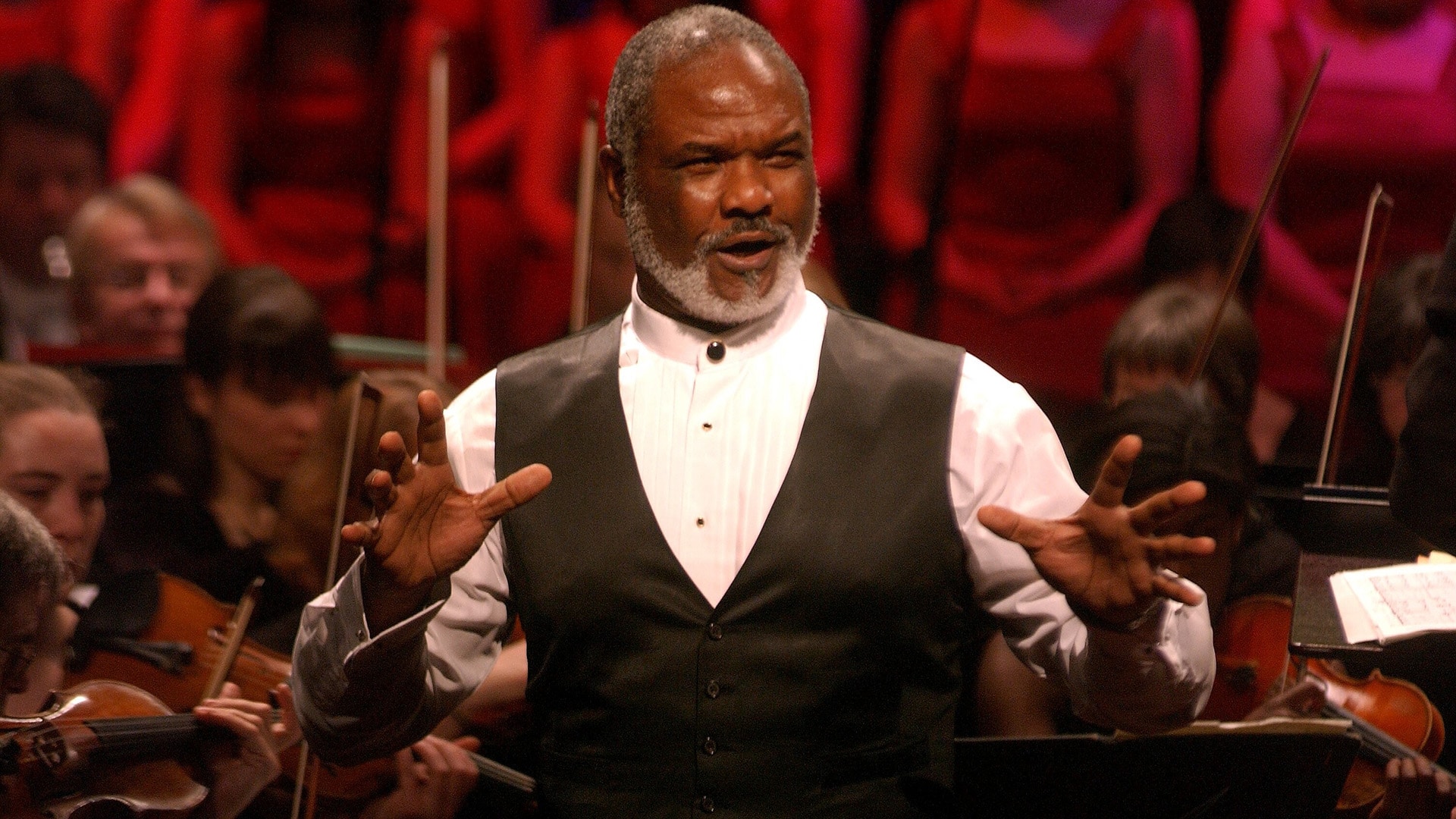 Willard White wearing a smart white shirt and black waistcoat, singing in front of an orchestra.