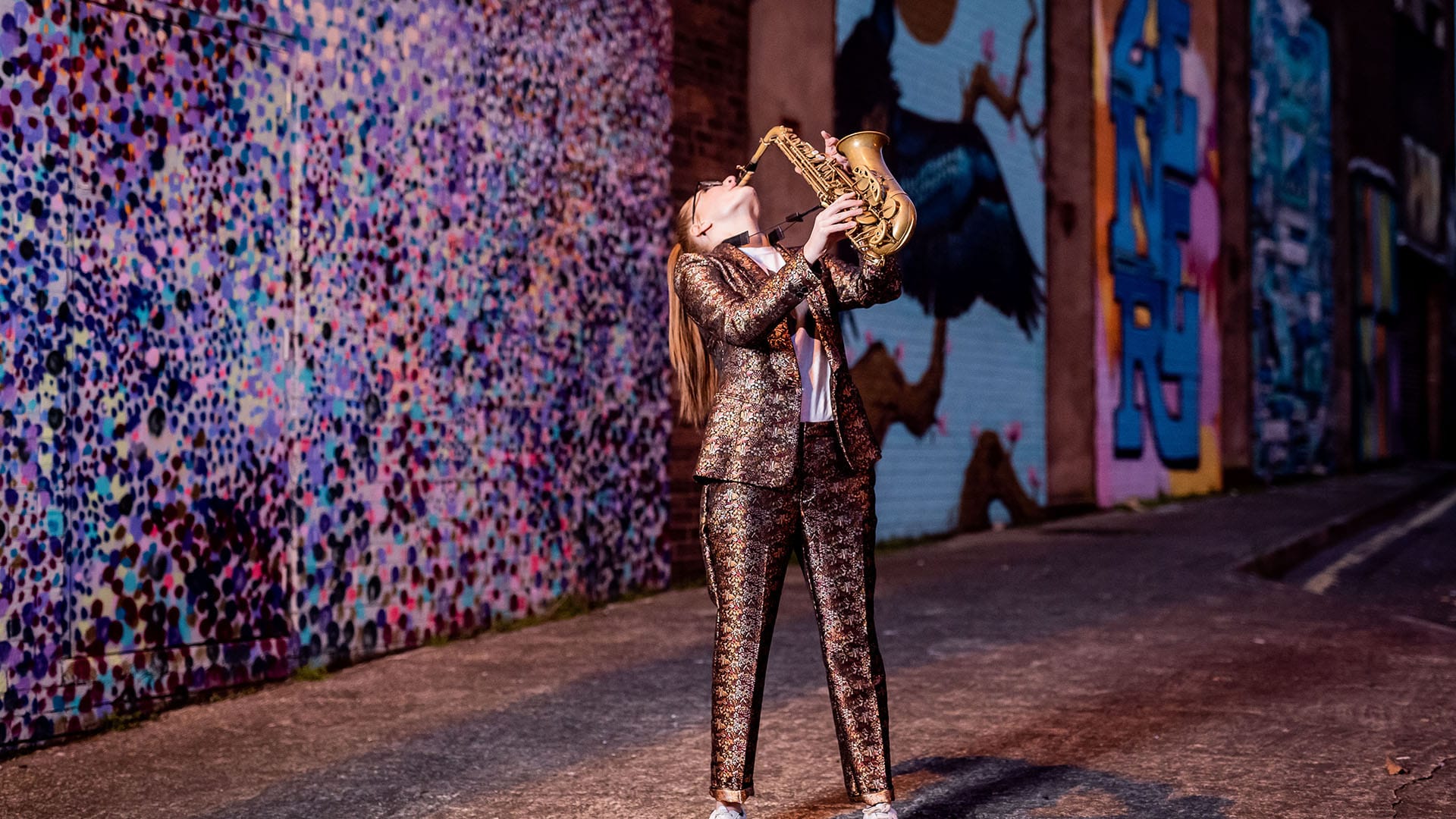 Saxophonist Jess Gillam stood outside, playing a saxophone, wearing a shiny colourful suit, with a long straight blonde ponytail.